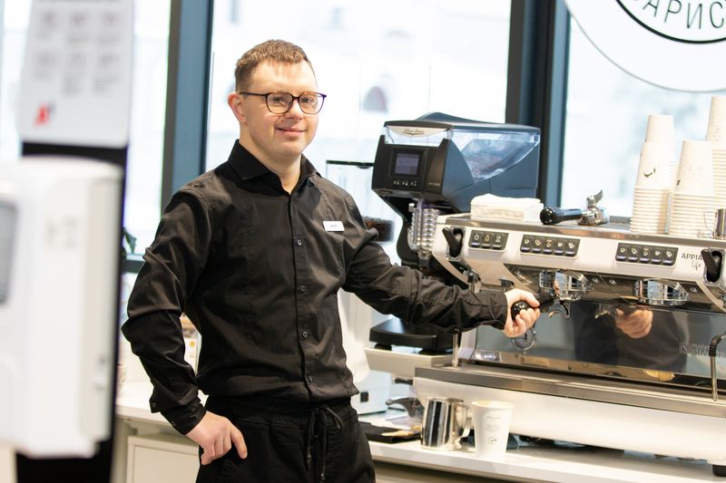 A barista in a black shirt stands proudly at the espresso machine in a cafe.