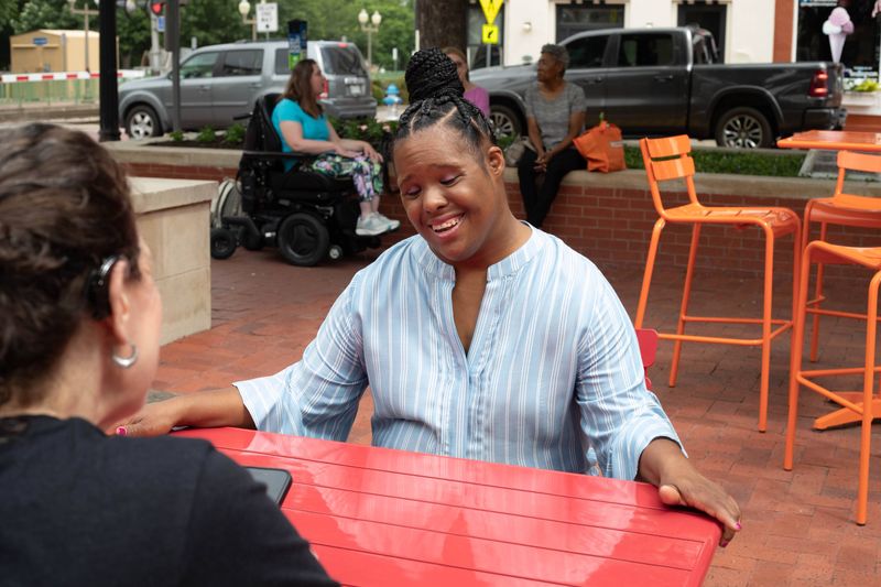 Two women talk and laugh across a red cafe table.