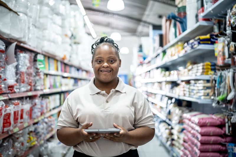 A smiling store worker stands in a grocery aisle holding a tablet.