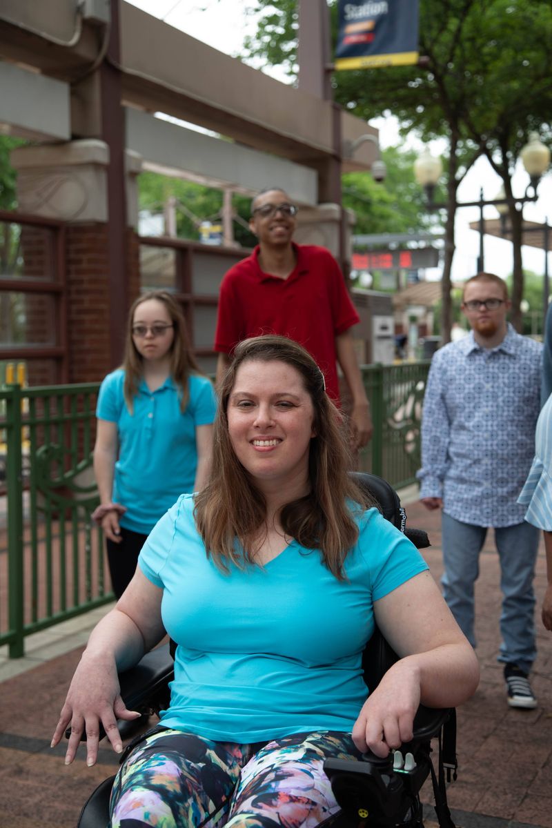A smiling woman in a power wheelchair with friends on a train platform.